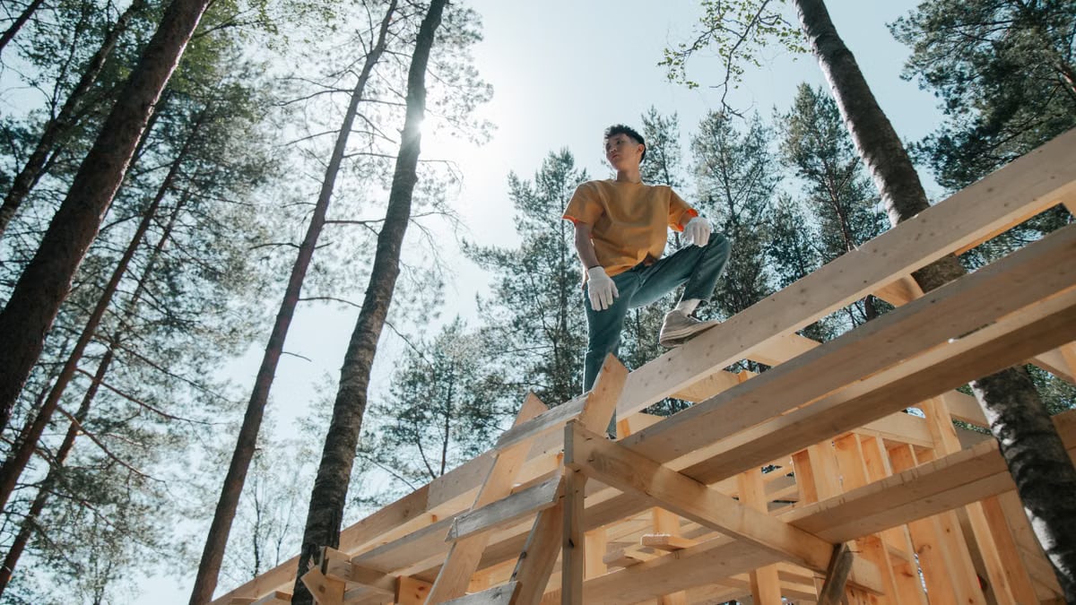 A carpenter on a wooden structure in a forest, engaged in construction work under the sun.